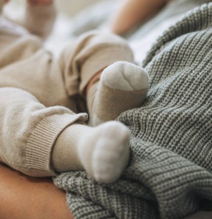 Cute baby feet in beige socks, child with mother on bed, happy motherhood